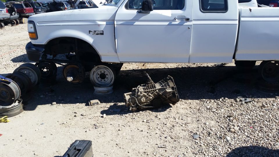 Transmission on ground next to a white Ford F-Series truck chassis missing wheels. Other vehicles in background