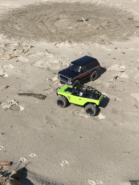 1989 Ford Bronco - Two remote control vehicles are parked on a sandy beach with some debris scattered around.