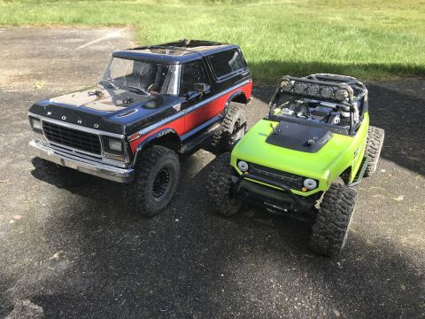 1989 Ford Bronco - Two RC vehicles, a black Bronco and a green Jeep, are parked on pavement with grass in the background.