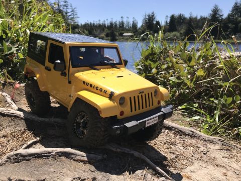 1989 Ford Bronco - Yellow Jeep Wrangler Rubicon on a dirt path with water and trees in the background.