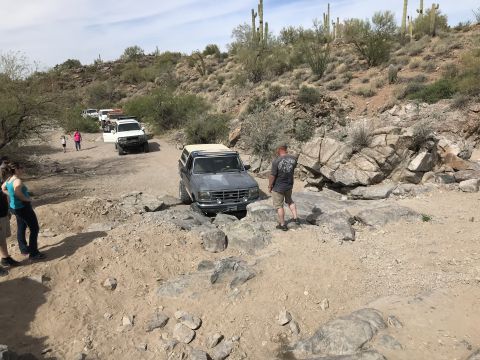 1991 Ford Bronco - Several off-road vehicles traverse a rocky desert trail under a partly cloudy sky.