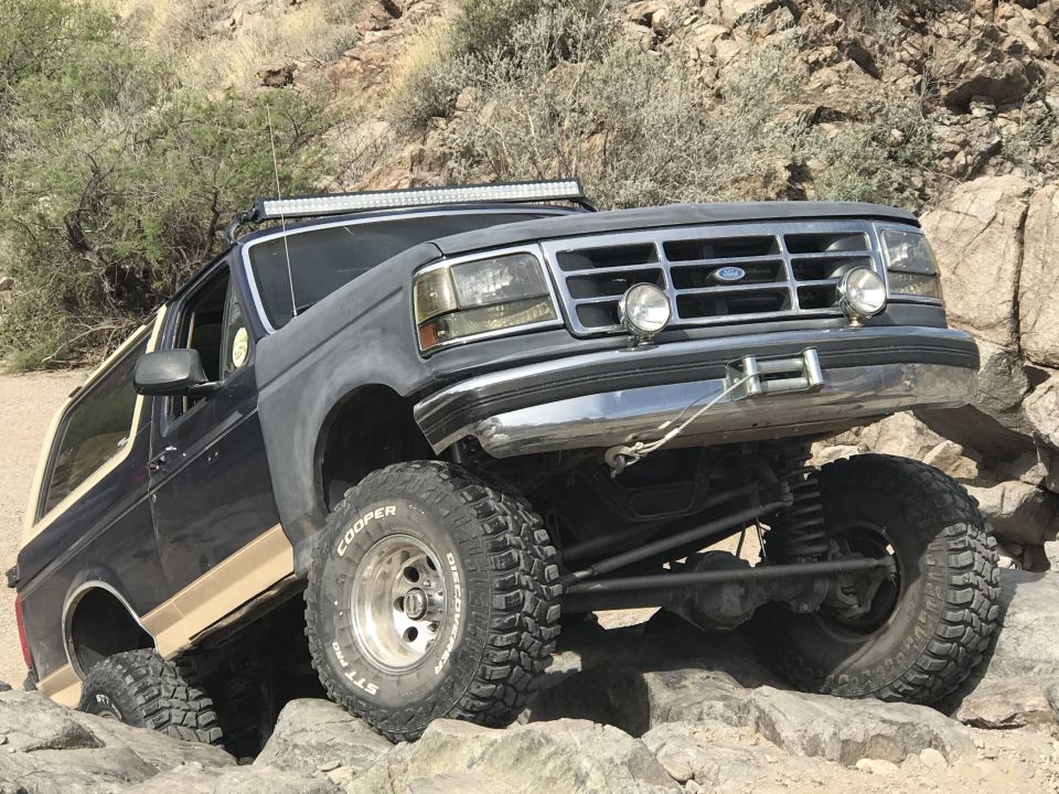 Dark gray Ford Bronco with tan lower body trim, lifted suspension, and off-road tires, navigating rocky terrain