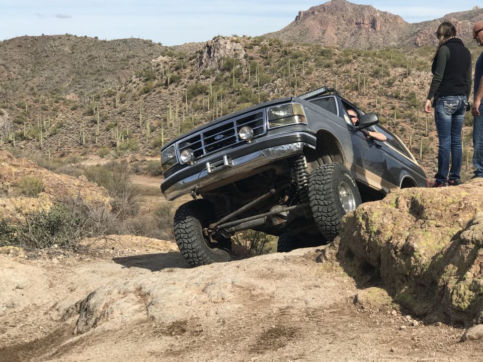 Ford Bronco climbing rocks on a desert trail, driver visible, two people watching