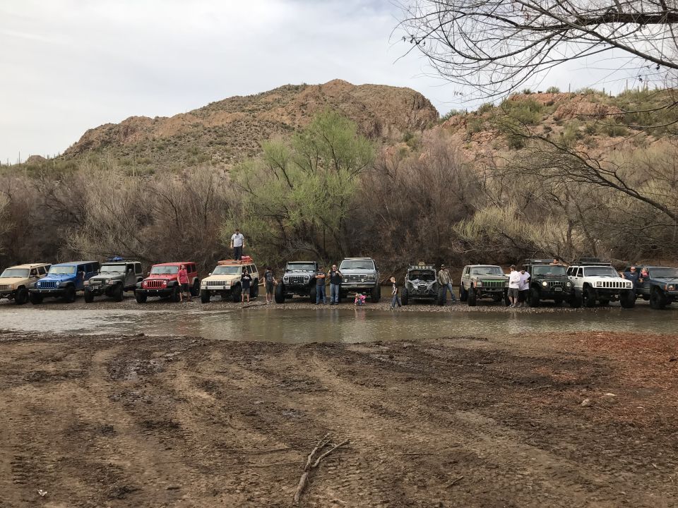 Multiple SUVs and off-road vehicles gathered in a muddy, shallow stream with a desert mountain in the background