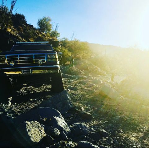 1991 Ford Bronco - A dark blue Ford Bronco navigates a rocky, off-road trail under a bright sun.
