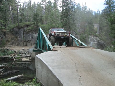 1968 Ford Bronco - Off-road SUV with large tires crosses a green metal bridge in a wooded area.
