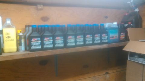 1992 Ford Bronco - A row of roughly a dozen black Amsoil synthetic motor oil quart bottles with blue caps lined up on a plywood garage shelf, alongside a blue…