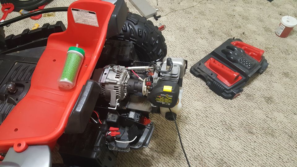 Disassembled red toy vehicle chassis with engine, alternator, and green glitter cup. Part of a black toy gun accessory is visible