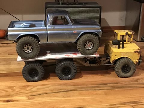 1989 Ford Bronco - A silver toy pickup truck is transported by a yellow toy truck with a six-wheel trailer.