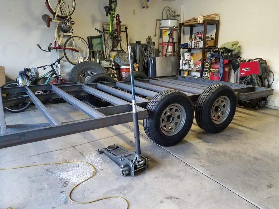 Custom trailer frame with two axles and tires in a garage, with a floor jack.