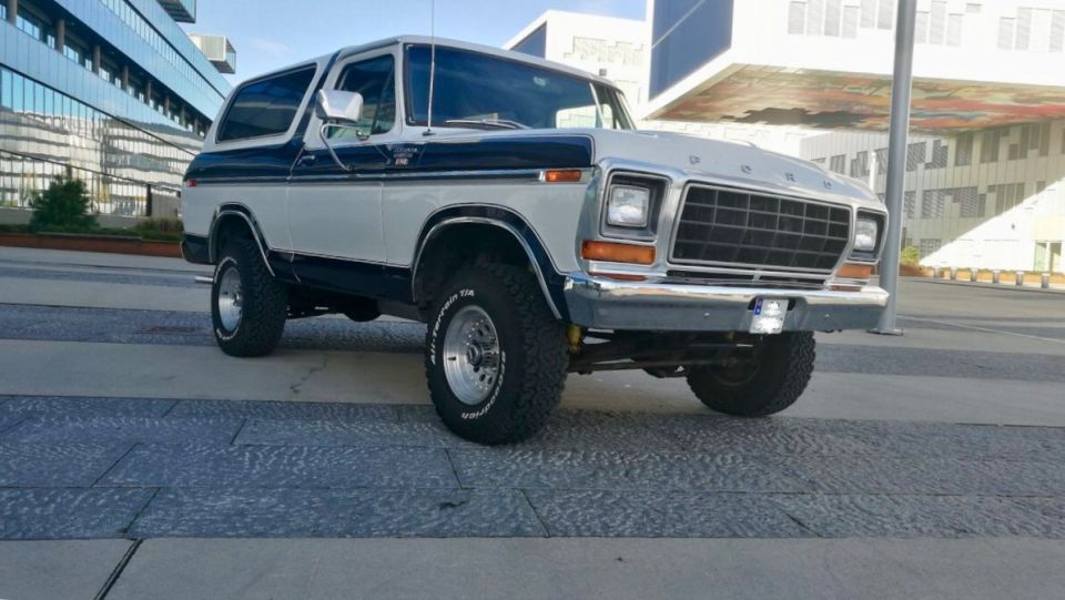1978 Ford Bronco, lifted, two-tone white and dark blue, parked in front of a modern building.