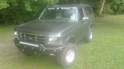 1992 Ford Bronco - Driver-side 3/4 front view of a lifted 1990s Ford Bronco in flat/satin dark paint parked on grass, with a black tubular front bumper carryi…