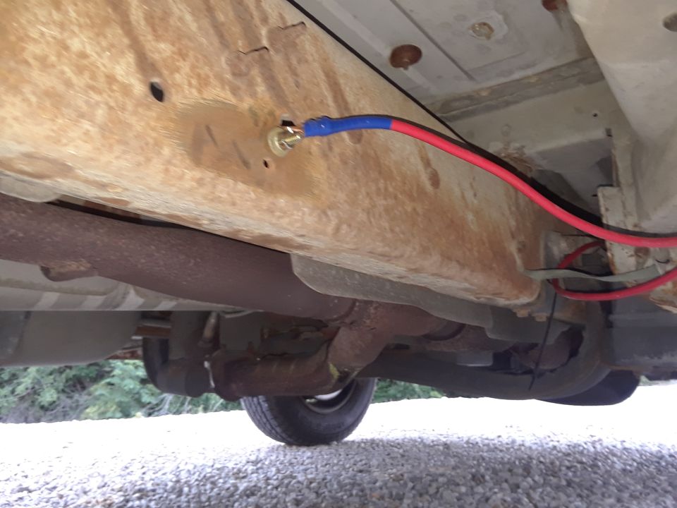 Underside of a rusty vehicle frame with red and blue wiring and exhaust pipes visible