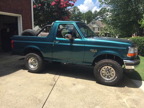 1996 Ford Bronco - Side view of a teal 1996 Ford Bronco with a spare tire mounted in the bed, parked outside a garage.