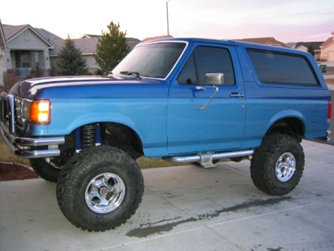 1989 Ford Bronco - This lifted blue Ford Bronco has large off-road tires and chrome wheels, with a chrome bumper and side steps.