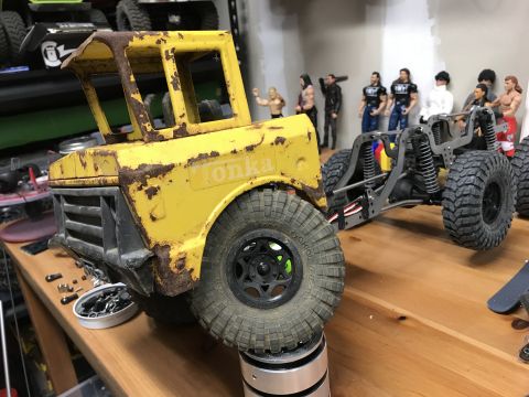 1989 Ford Bronco - A weathered yellow Tonka truck body sits on a stand, revealing its chassis and large off-road tires, with toy figures in the background.