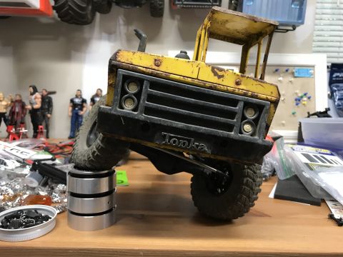 1989 Ford Bronco - A weathered yellow toy truck sits on a workbench, its front wheel elevated on a metal stand.