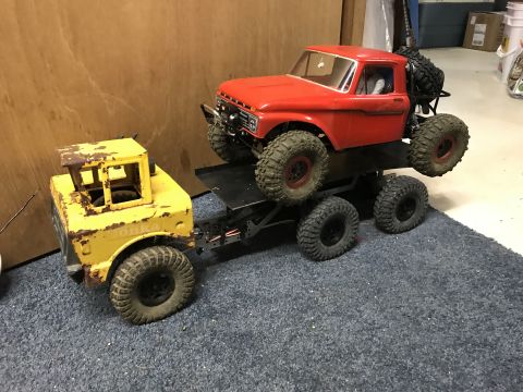 1989 Ford Bronco - A red pickup truck is being towed by a yellow, six-wheeled tow truck, both appearing to be RC models.