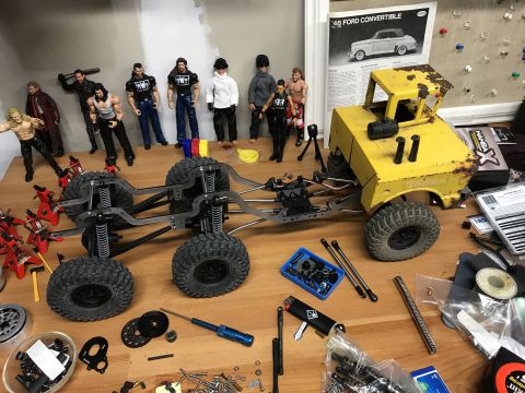 1989 Ford Bronco - A collection of RC car parts, action figures, and tools are laid out on a wooden surface, suggesting a hobbyist's workspace.
