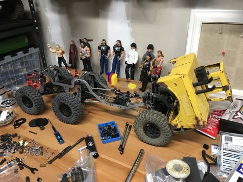 1989 Ford Bronco - Two scale RC rock-crawler chassis under build on a workbench, lined with WWE wrestler action figures and scattered tools.