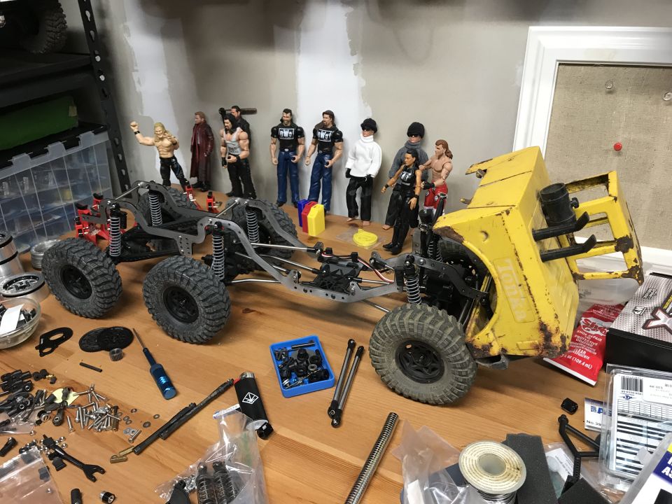 RC crawler chassis builds on workbench with row of wrestling action figures