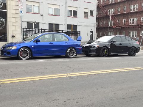 2006 Mitsubishi Evolution - A blue Mitsubishi Lancer Evolution IX and a black Subaru WRX are parked side-by-side on a city street.