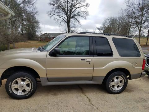 2003 Ford Explorer - A tan 2003 Ford Explorer is shown from the side, parked on a concrete driveway with a house and trees in the background.