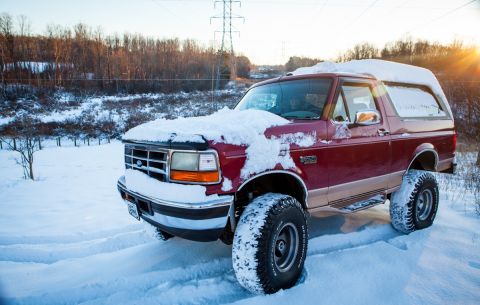 1995 Ford Bronco - A red Ford Bronco sits partially covered in snow on a winter day, with the sun setting behind bare trees.