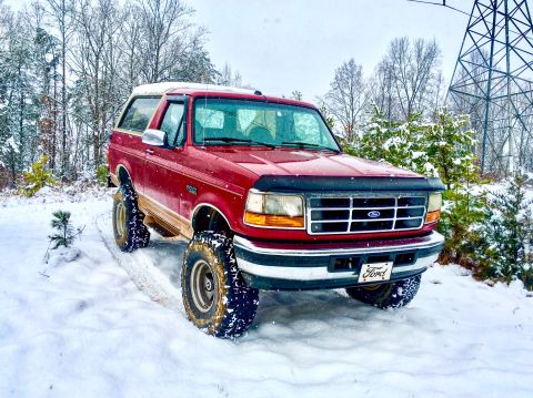 1995 Ford Bronco - A red 1995 Ford Bronco equipped with large off-road tires navigates a snowy landscape.