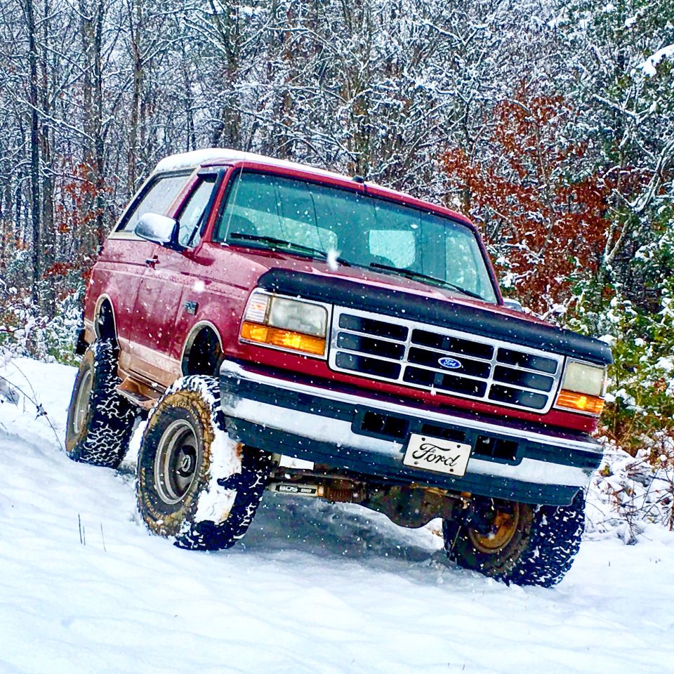 Red 1995 Ford Bronco driving through snow with trees in the background