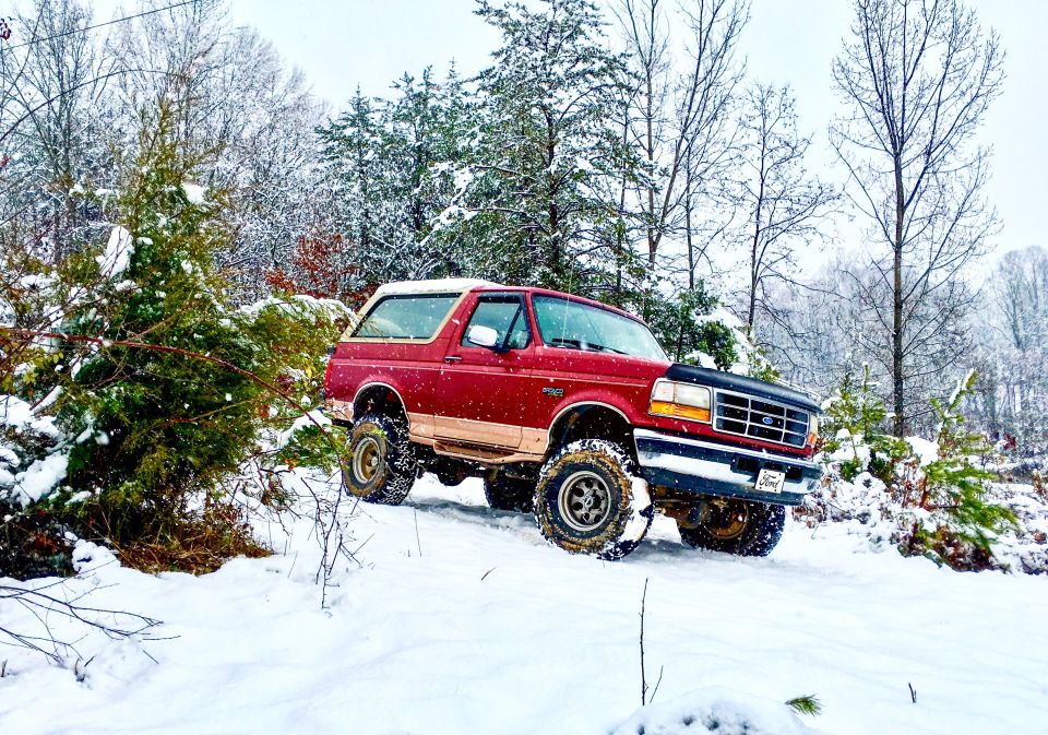 Red Ford Bronco with tan trim driving on a snowy trail, front 3/4 view