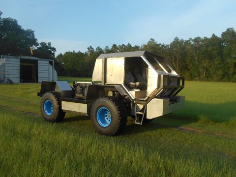 1992 Ford Bronco - A custom-built, four-wheeled vehicle with a silver, angular body and large, off-road tires with blue rims sits in a grassy field. A metal b…
