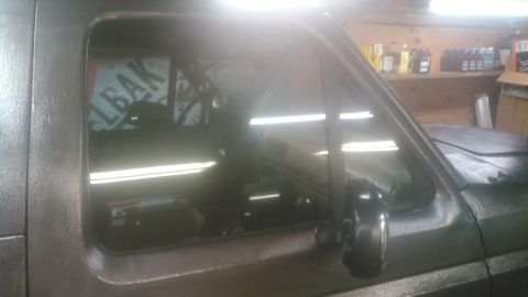 1992 Ford Bronco - Close view of the driver's door glass and vent window on a flat-gray truck inside a garage, with shop lights reflecting off the glass and a…
