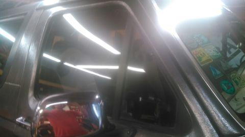 1992 Ford Bronco - Close-up of a truck's driver-side door glass and vent window from outside in a garage, with shop lighting reflecting in long streaks off th…