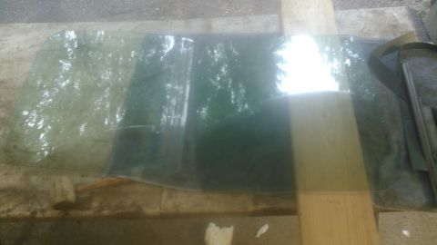 1992 Ford Bronco - A large, curved piece of green tinted glass, likely a windshield or rear window, rests on a wooden surface outdoors. The glass reflects tre…
