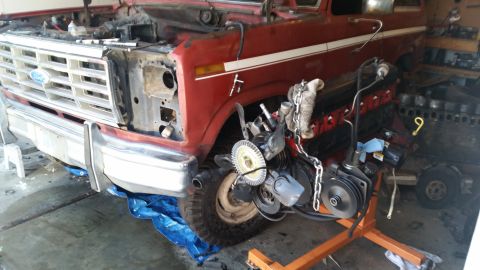 1981 Ford Bronco - A red 1980s Ford truck is undergoing engine work, with the engine partially removed and resting on an engine stand. The front grille, bumpe…