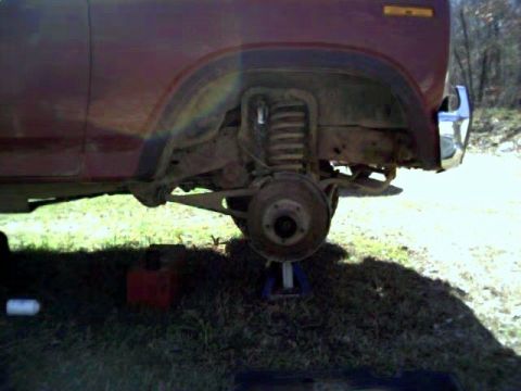 1986 Ford Bronco - The front wheel well and suspension of a red SUV are shown, with the vehicle supported by a jack.