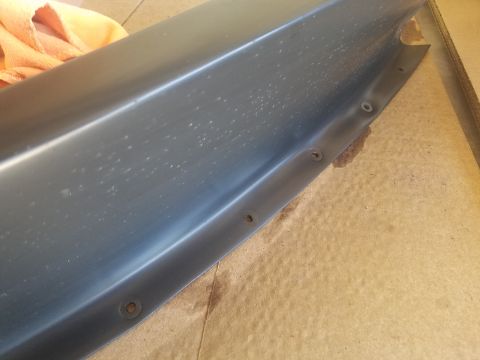 1968 Ford Bronco - A close-up view of a dark gray, possibly metal, vehicle part with visible rivets along the edge. The part appears to be curved and is resti…