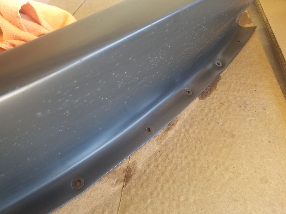 Close-up of a dark gray vehicle part with rivets, resting on a brown surface.