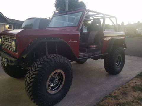 1968 Ford Bronco - A customized red 1968 Ford Bronco is parked on a concrete driveway in front of a house. The vehicle features a roll cage, large off-road ti…