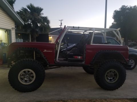 1968 Ford Bronco - The side profile of a modified red 1968 Ford Bronco is shown parked on a concrete driveway. The vehicle features a white roll cage, large o…