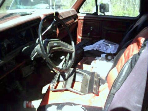 1986 Ford Bronco - The interior of a red 1980s Ford truck is visible, with a disassembled dashboard piece on the passenger seat.