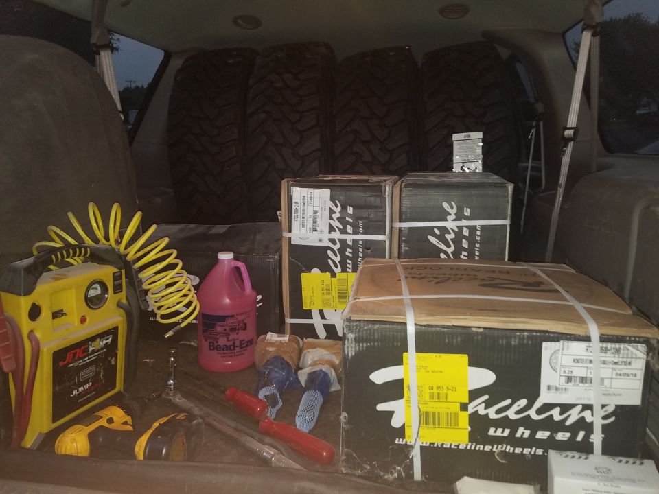 Four large tires and three boxes of Raceline wheels in the cargo area of a vehicle with tools.