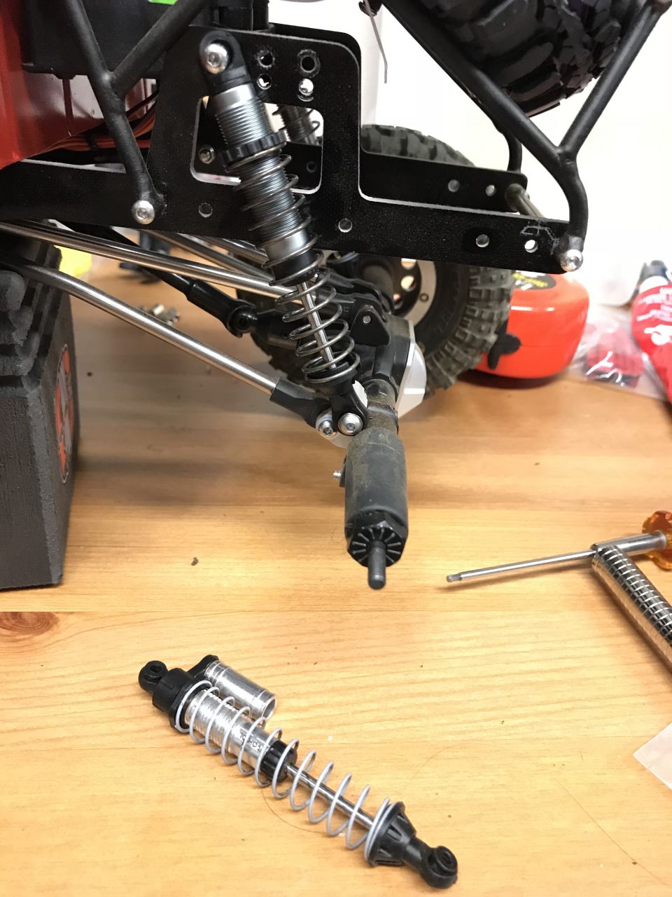Disassembled RC car shock absorber on a wooden table, with vehicle parts in the background.
