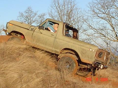 1978 Ford Bronco - A mud-caked olive-green full-size Ford Bronco with the top removed climbs a steep, dry grass hillside, body angled nose-up against a blue s…