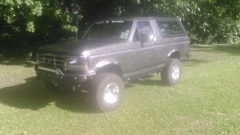 1992 Ford Bronco - Three-quarter front view of a lifted gray Ford Bronco parked on grass in the shade, with freshly tinted side and rear glass and a white 'Br…