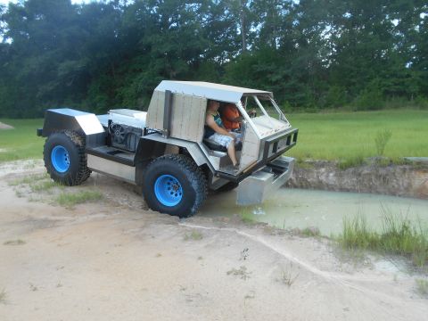1992 Ford Bronco - A custom-built, amphibious-looking vehicle with large tires and a metal body is driving through a muddy ditch. Two people are visible insid…