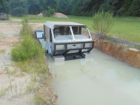 1992 Ford Bronco - A custom-built, amphibious vehicle with a silver, boxy cabin is partially submerged in a muddy trench. The vehicle has a large, exposed fro…