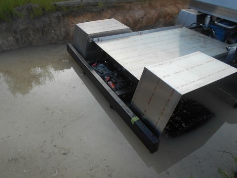 1992 Ford Bronco - A partially submerged custom-built aluminum watercraft with a visible battery and engine components sits in muddy water next to a dirt emba…
