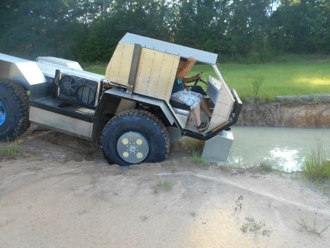 1992 Ford Bronco - A custom-built, amphibious vehicle with large tires is partially submerged in muddy water, with a person driving it from an open-air cabin.